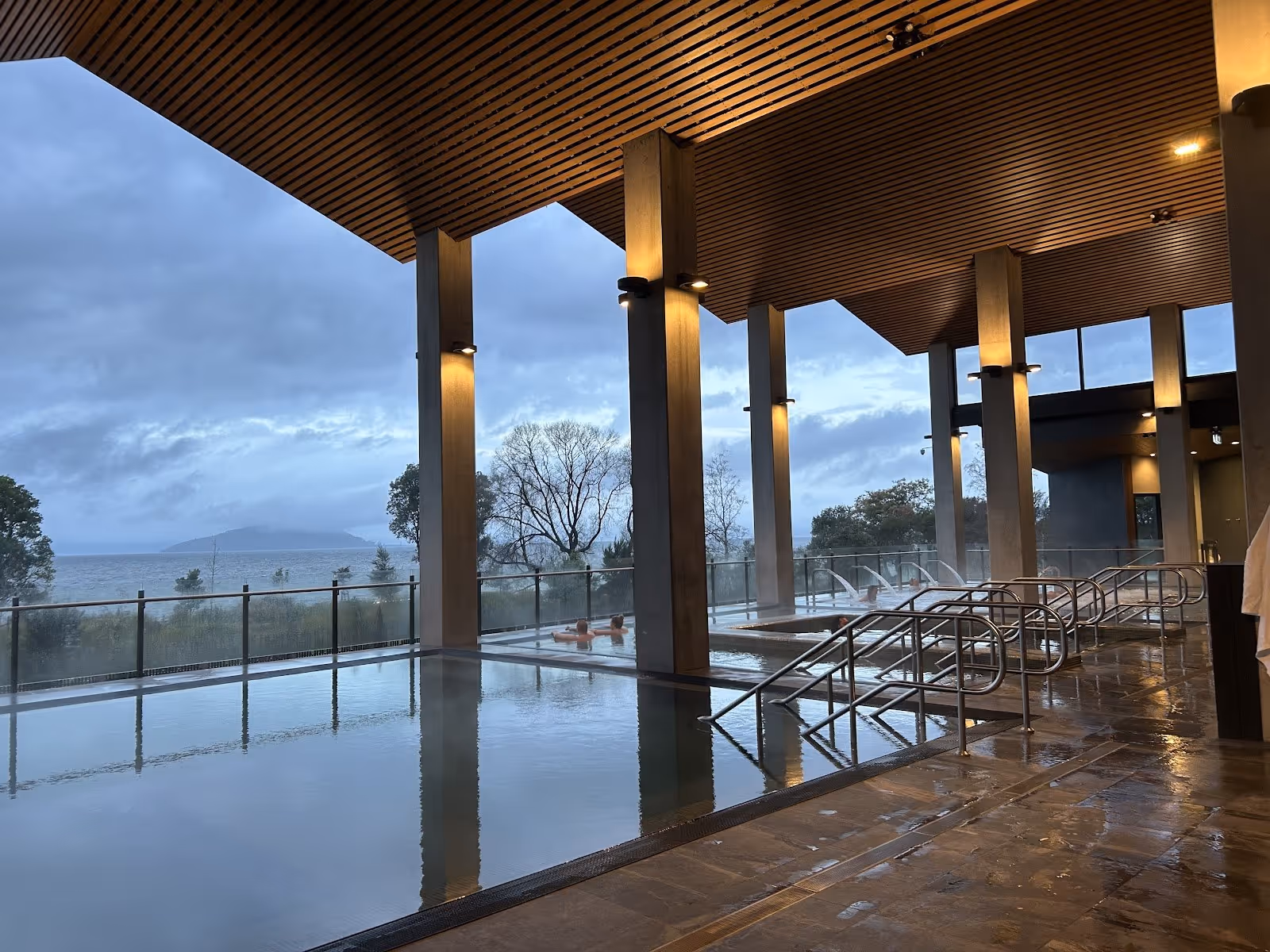 Multiple steaming hot spring soaking pools at Wai Ariki with metal handrails and glass fencing facing a lake and cloudy sky in Rotorua New.
