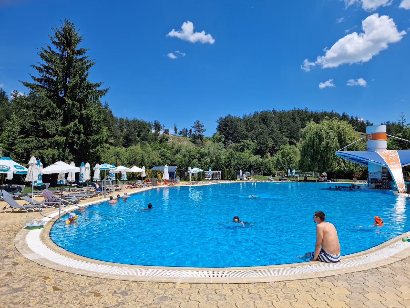 Wide view of a circular outdoor soaking pool at Alpha Spa & Pool surrounded by green trees under a clear sky in Bulgaria.