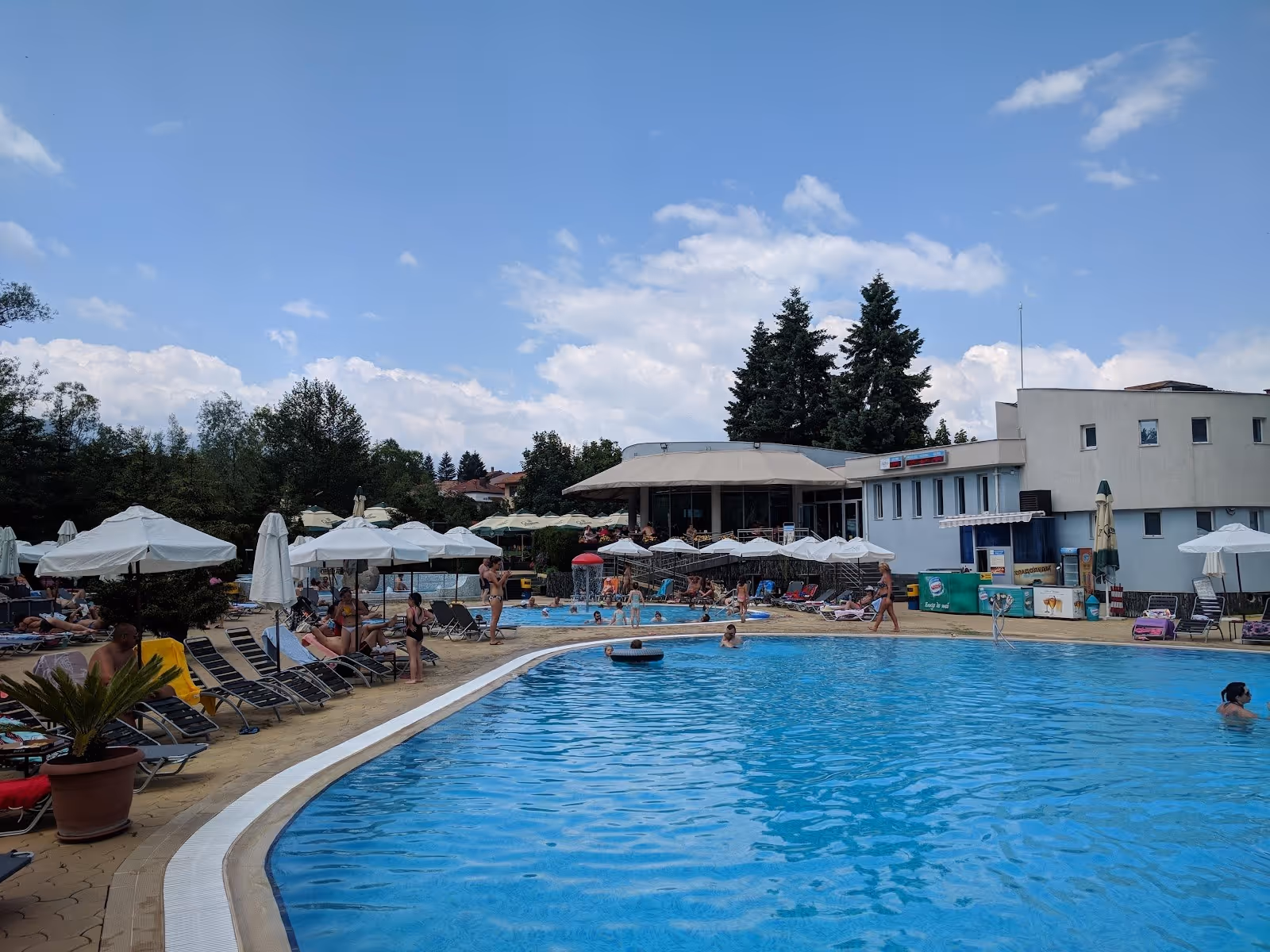 People relaxing and swimming in outdoor hot spring pools near a white building at Alpha Spa & Pool in Dobrinishte, Bulgaria.