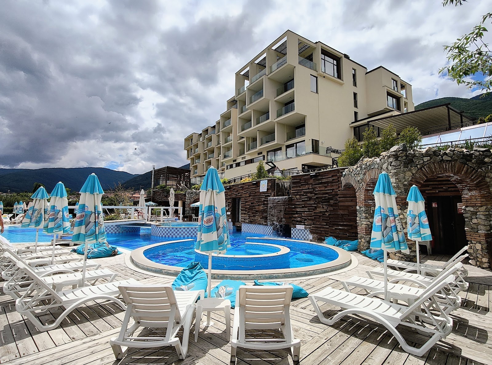Blue-tiled circular mineral pool surrounded by lounge chairs and umbrellas at Aqua Club Kotvata with resort building in Sapareva Banya.