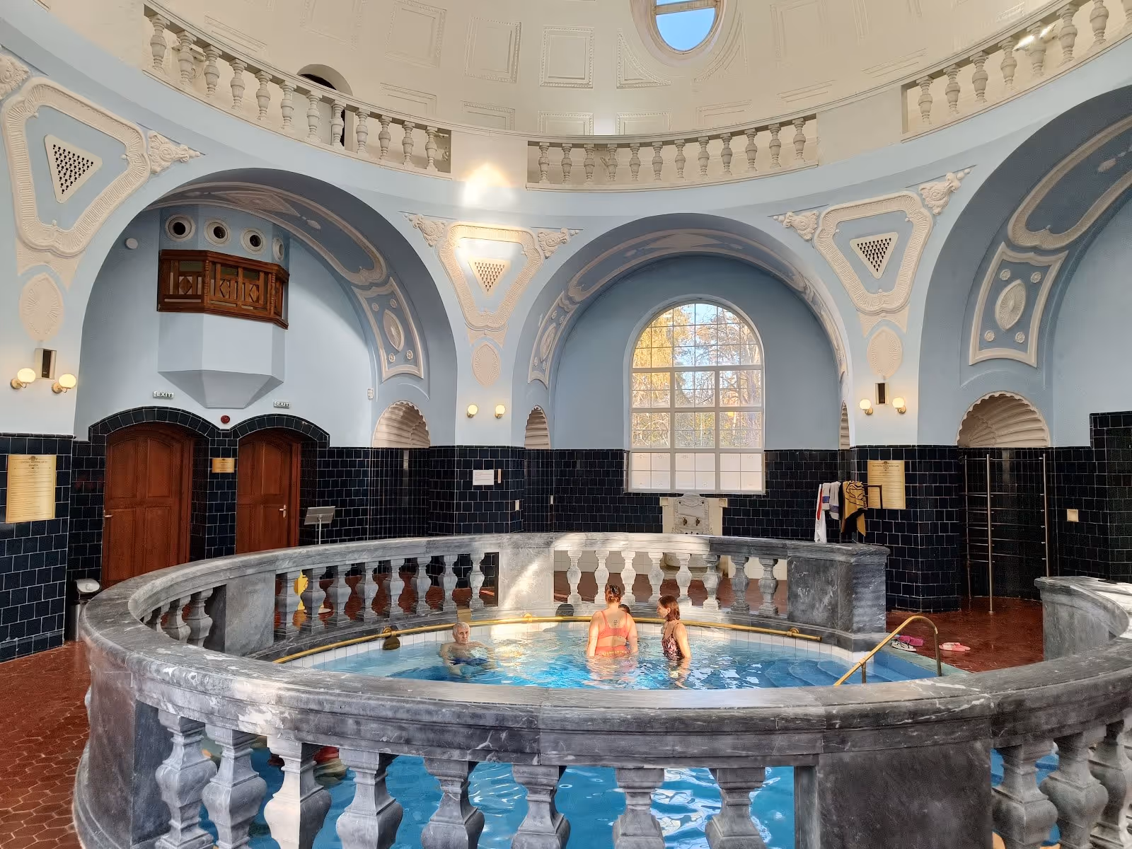 People relaxing in the indoor thermal pool with light blue walls and a large rounded window at Central Mineral Bath Bankya near Sofia Bulga.