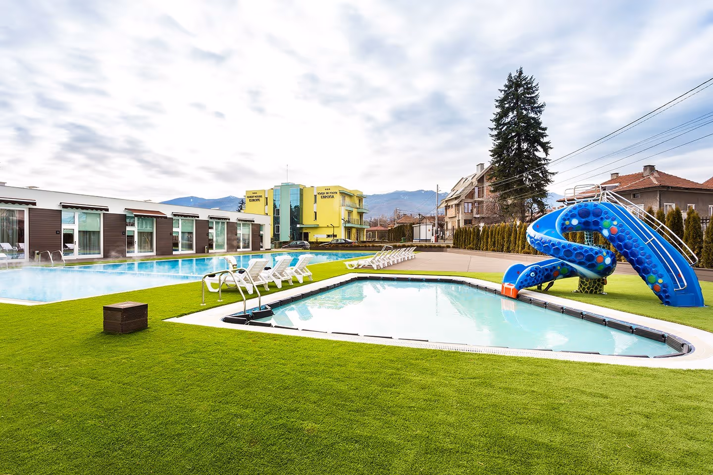 Outdoor mineral pools with steam rising, surrounded by white lounge chairs and a nearby colorful water slide at Complex Europe Mineral Pool.