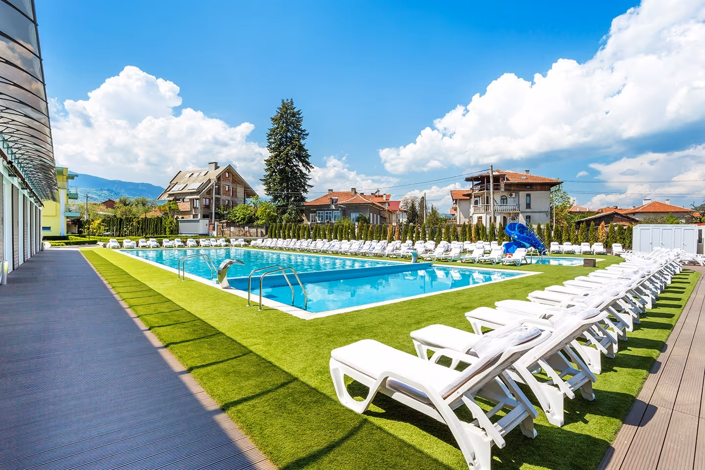 Large rectangular mineral pool bordered by rows of white lounge chairs and green grass at Complex Europe Mineral Pool with houses and mount.