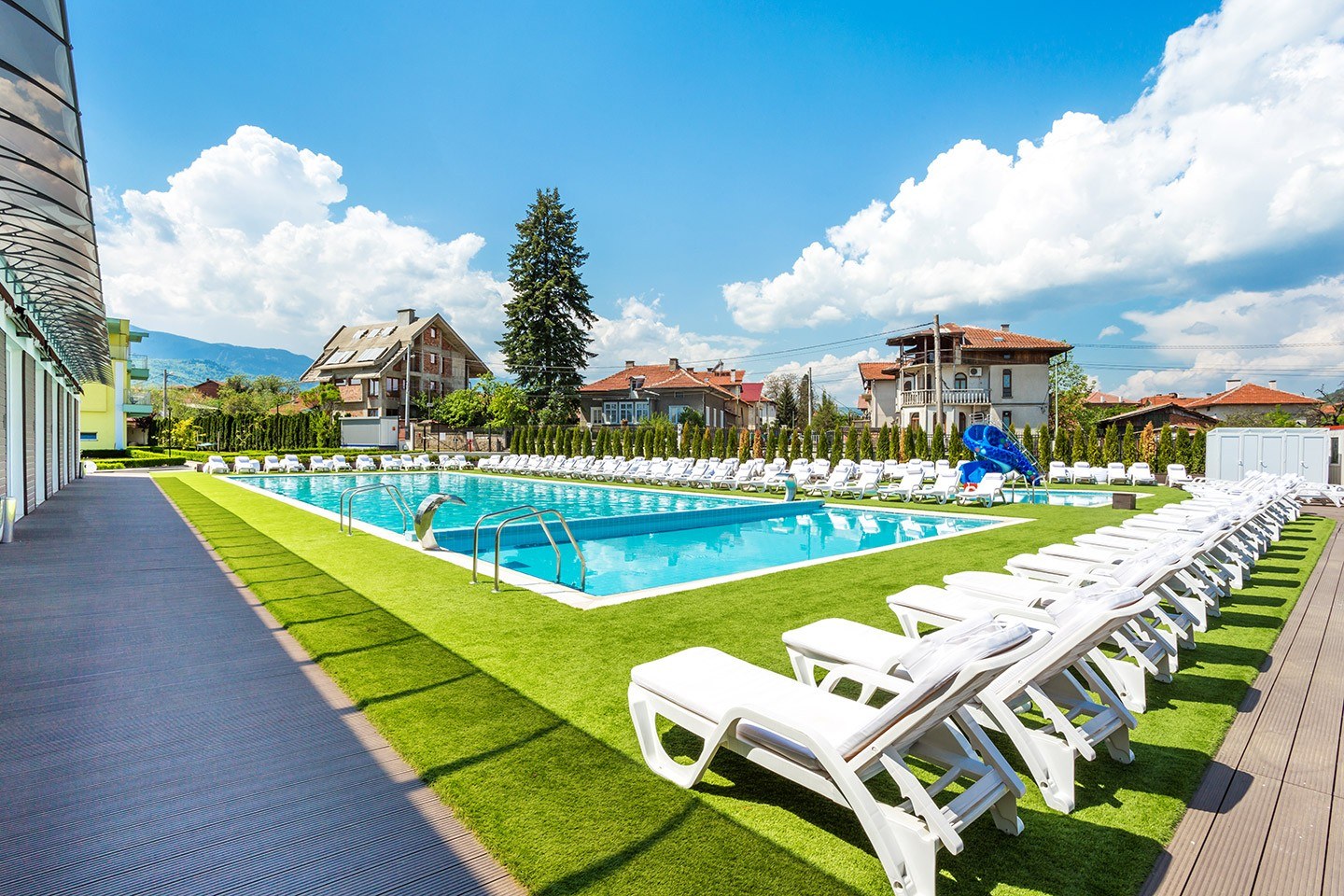 Large rectangular mineral pool bordered by rows of white lounge chairs and green grass at Complex Europe Mineral Pool with houses and mount.