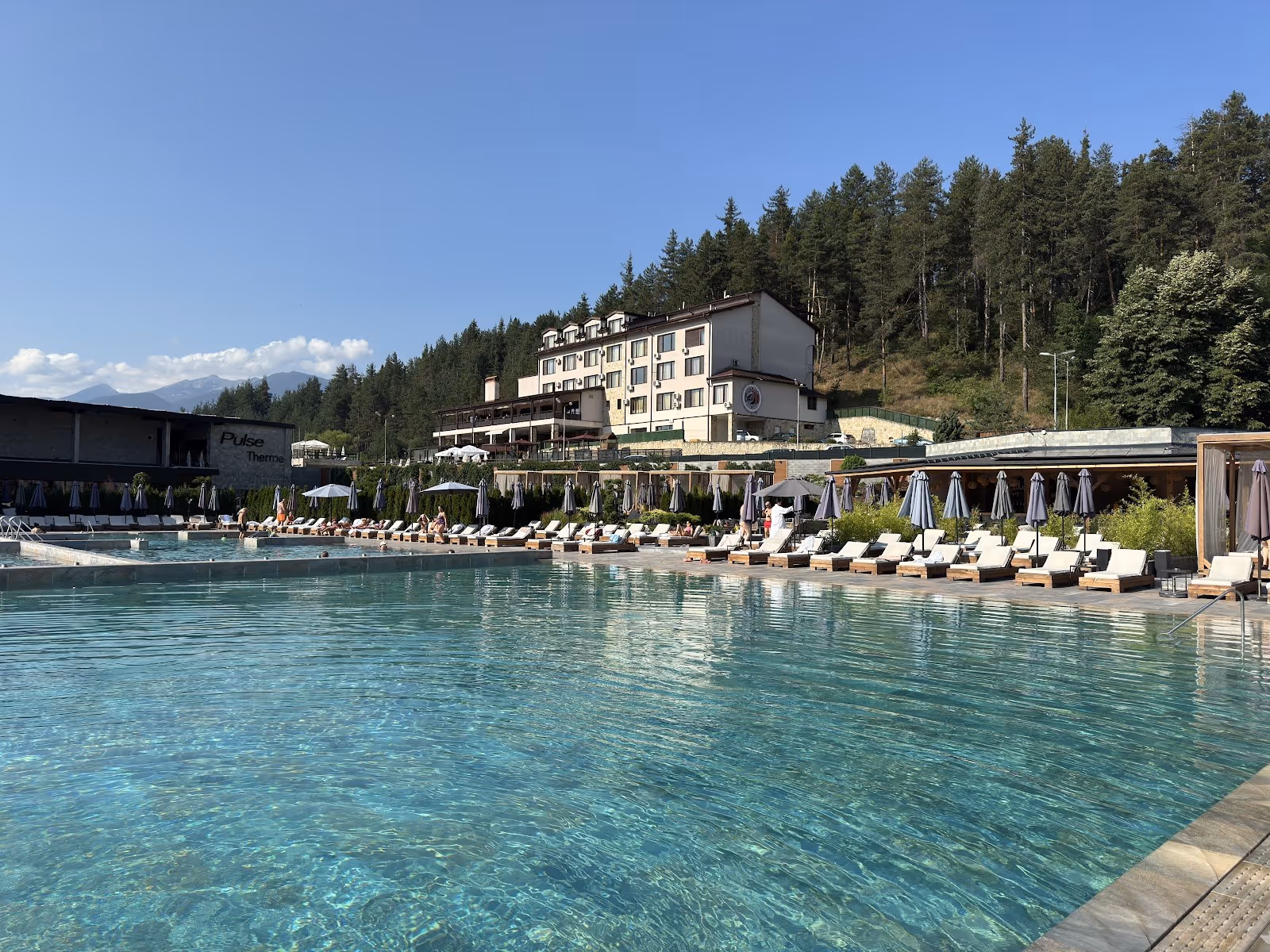 Large outdoor pool with clear water and numerous sun chairs by the edge, surrounded by pine trees and mountains in Banya near Bansko, Bulga.