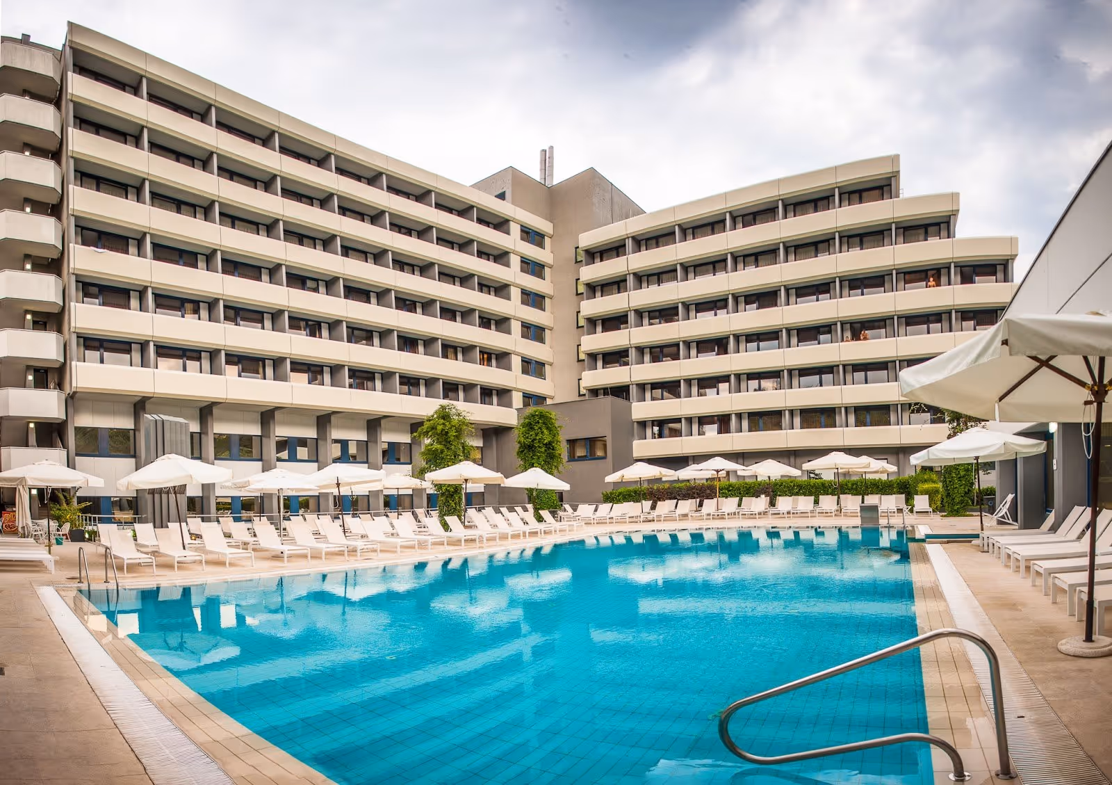 Large outdoor mineral pool surrounded by white umbrellas and lounge chairs with a modern hotel building at Interhotel Sandanski Thermal Poo.