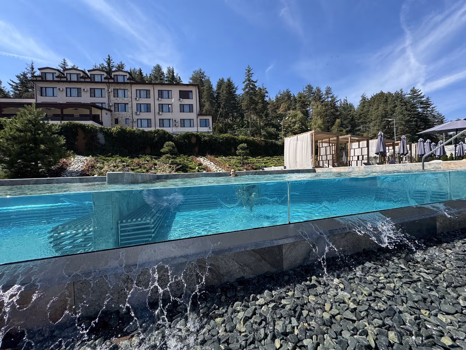Outdoor transparent-walled hot spring pool at Pulse Therme Banya showing water cascading over stones with trees and a white building nearby.