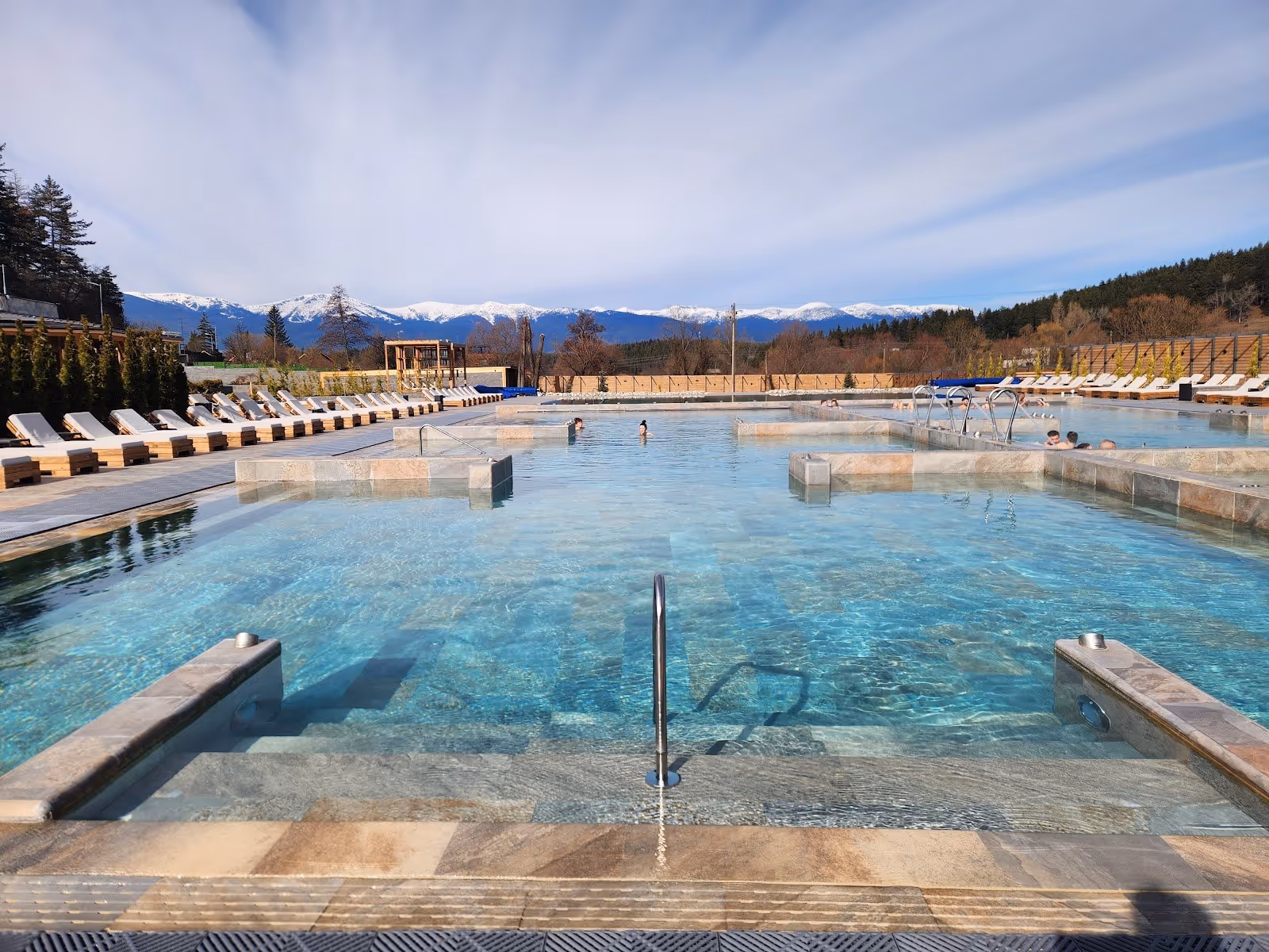 Outdoor hot spring pool at Pulse Therme Banya with clear water and lounge chairs, snow-capped mountains in the background near Banya, Bulga.