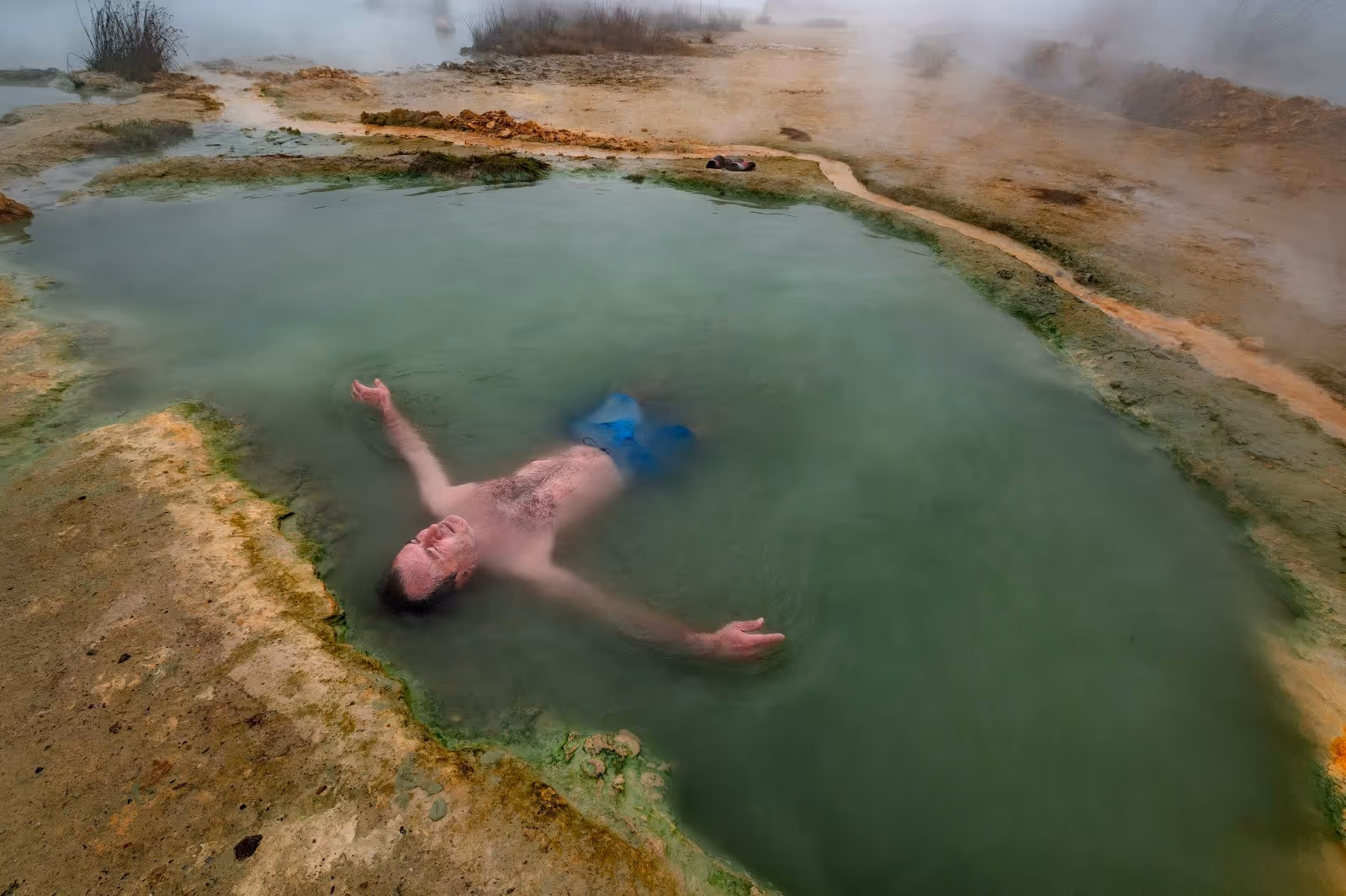 A man relaxing in a small, green mineral hot spring pool with steam rising at Rupite Thermal Springs near Petrich Bulgaria.