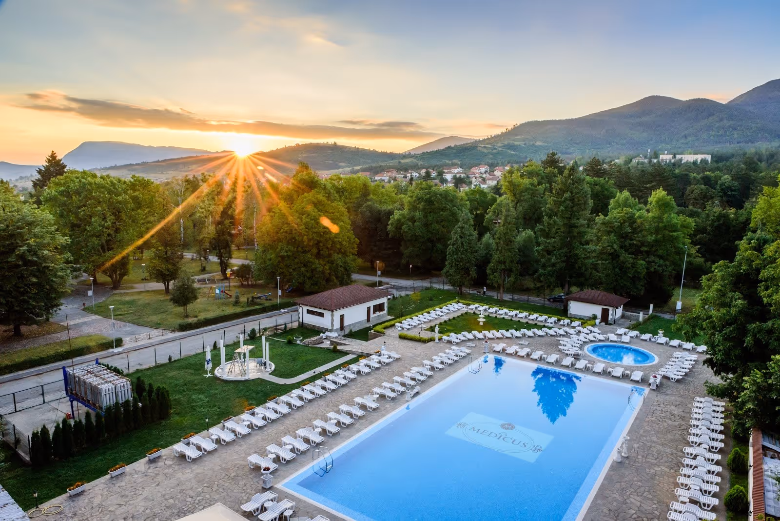 Outdoor hot spring pool of SPA Hotel Medicus with empty white lounge chairs and sunset behind hills near Varshets Bulgaria.