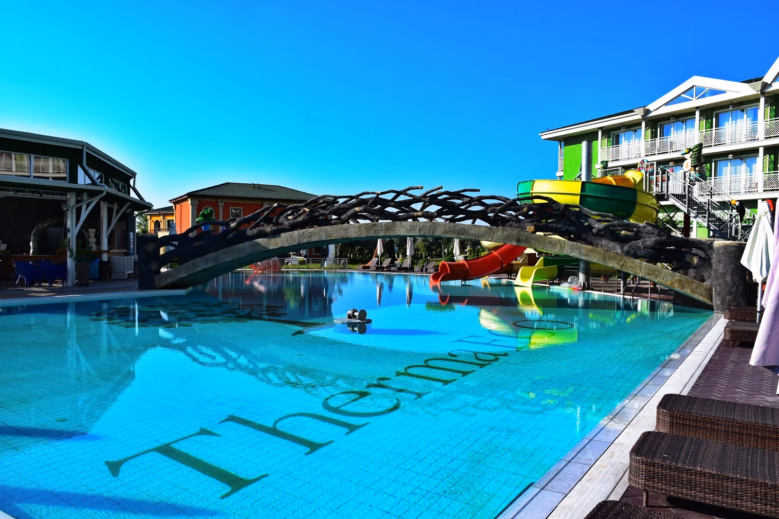 Outdoor mineral pool at Therma ECO Village Mineral Pools in Varna Bulgaria with a wooden bridge and water slides under clear blue sky.