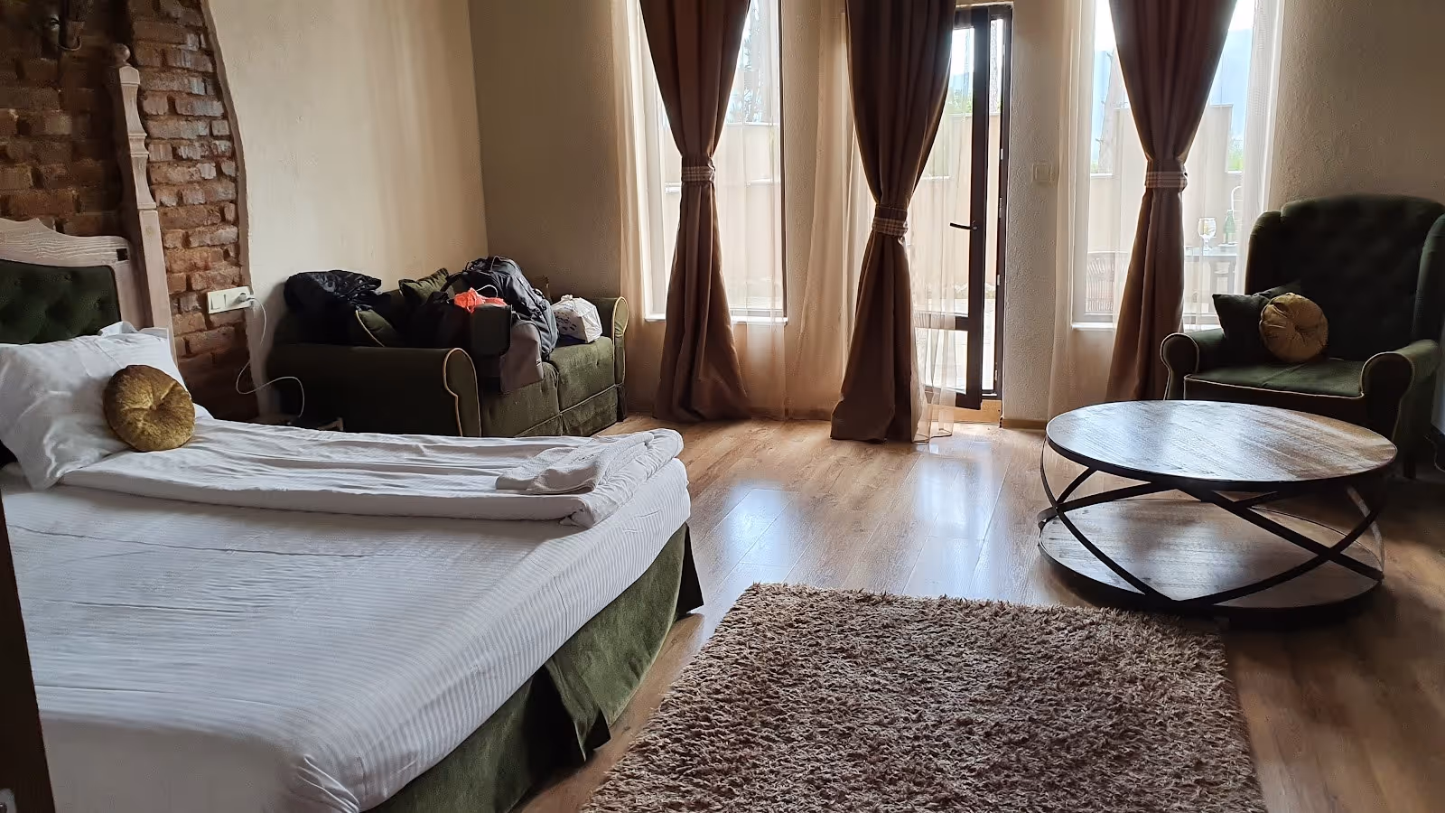 Interior room with green velvet armchairs, a bed with white linens, large windows with sheer curtains, and wooden floor.