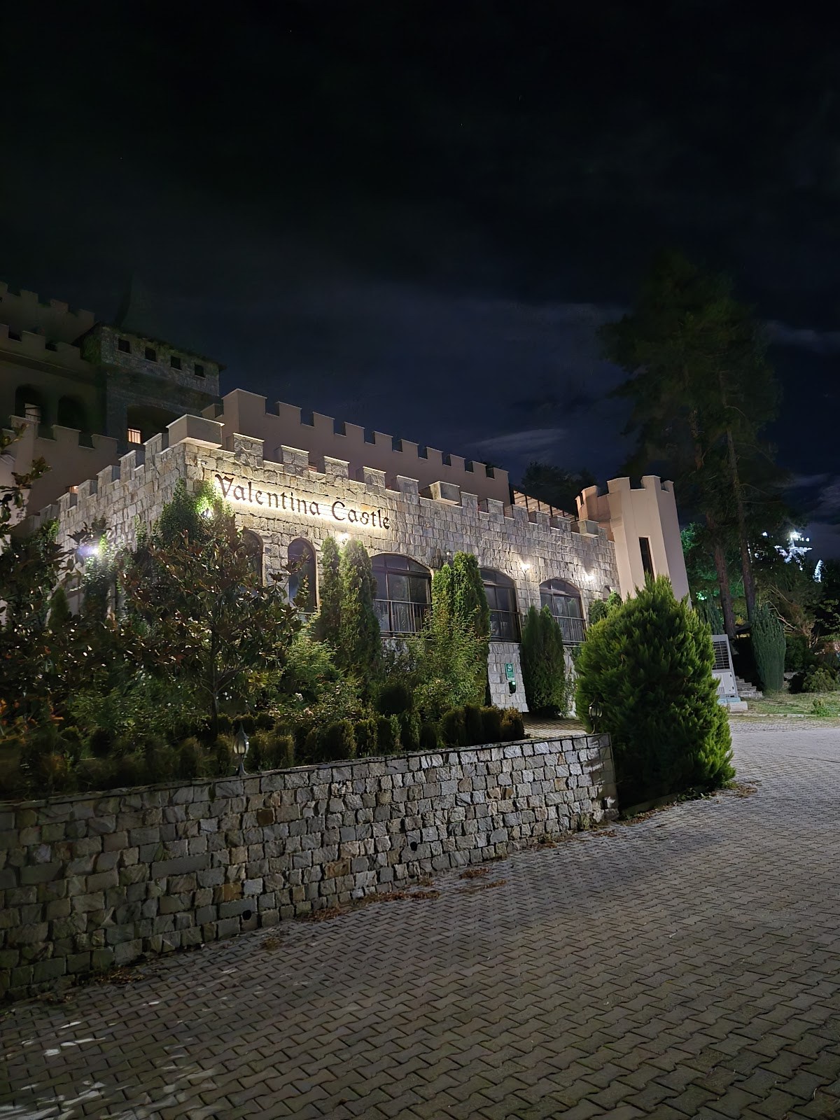 Stone castle building with Valentina Castle sign illuminated at night nestled among pine trees near Ognyanovo Bulgaria.