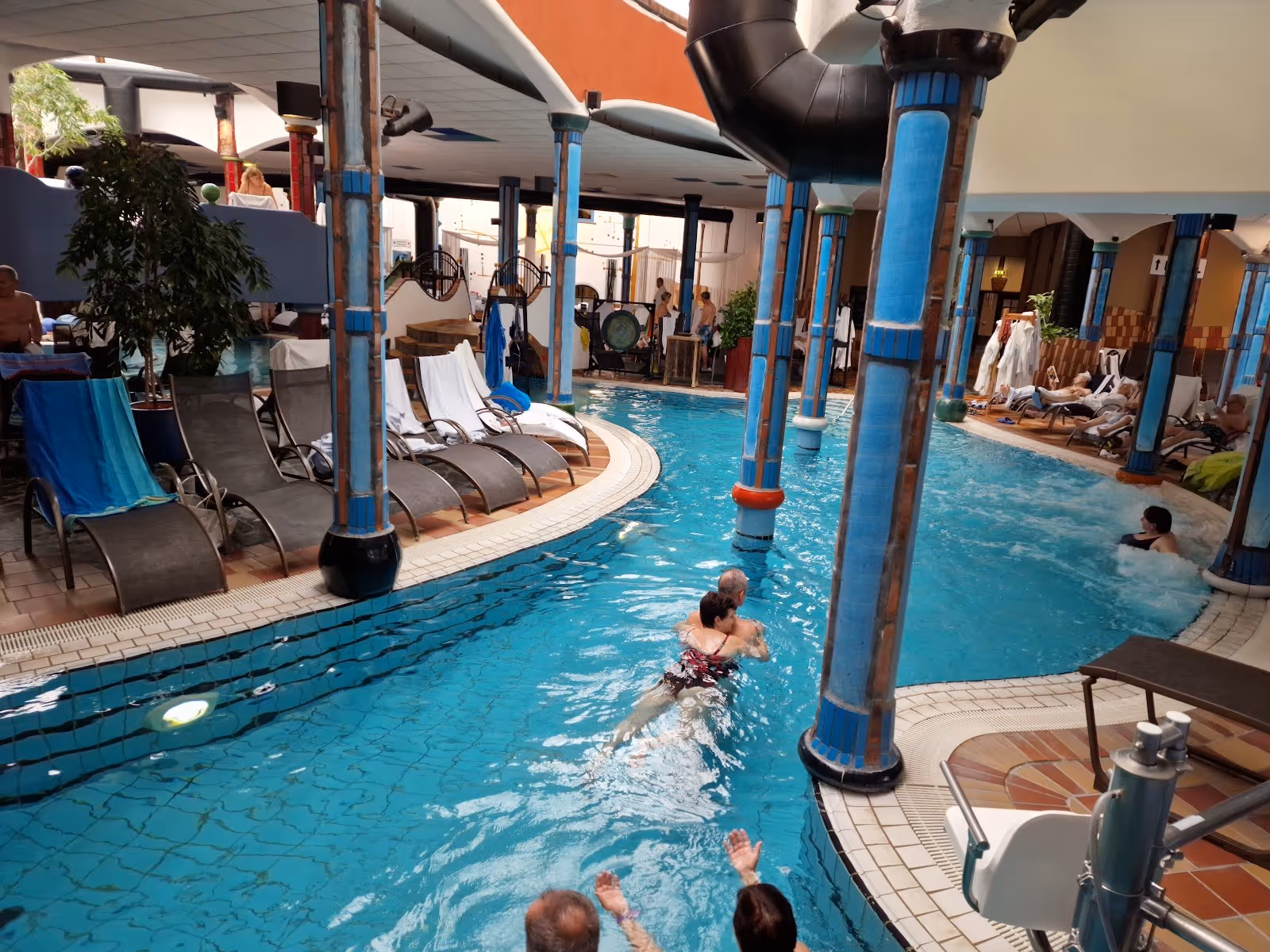Indoor hot spring pool surrounded by tall blue columns and lounge chairs with several people relaxing in the water at Rogner Bad Blumau Aus.