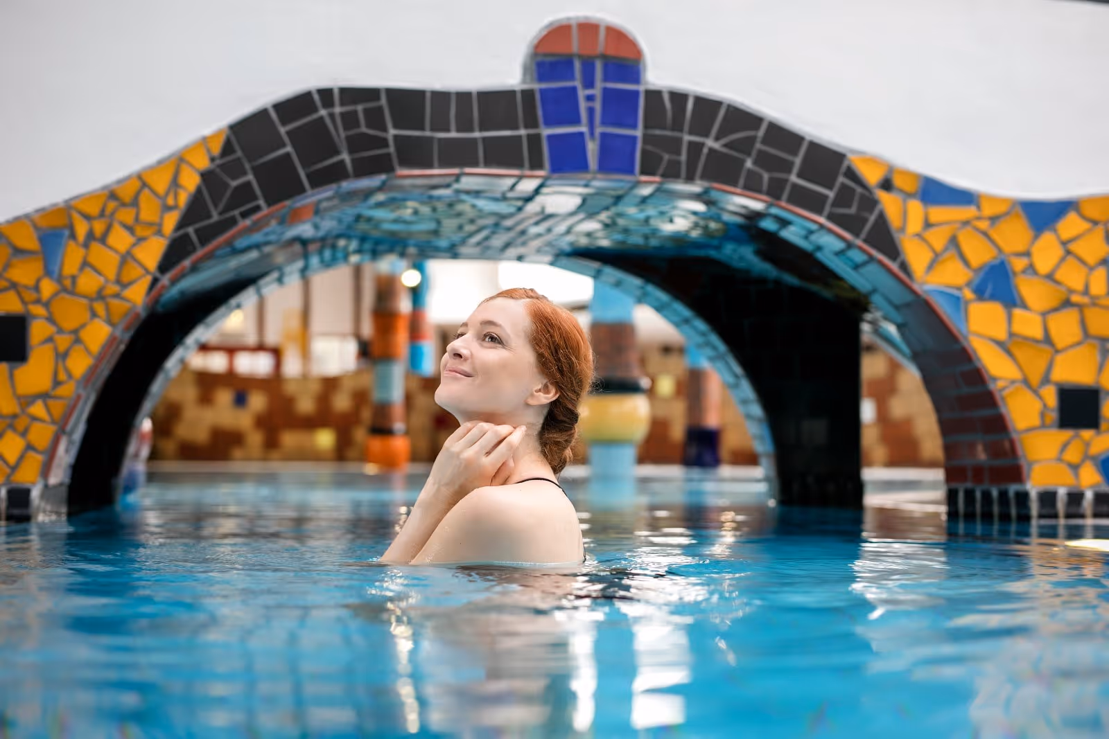 Red-haired woman enjoying the blue-tiled indoor mineral pool beneath a colorful mosaic archway at Rogner Bad Blumau hot spring in Austria.