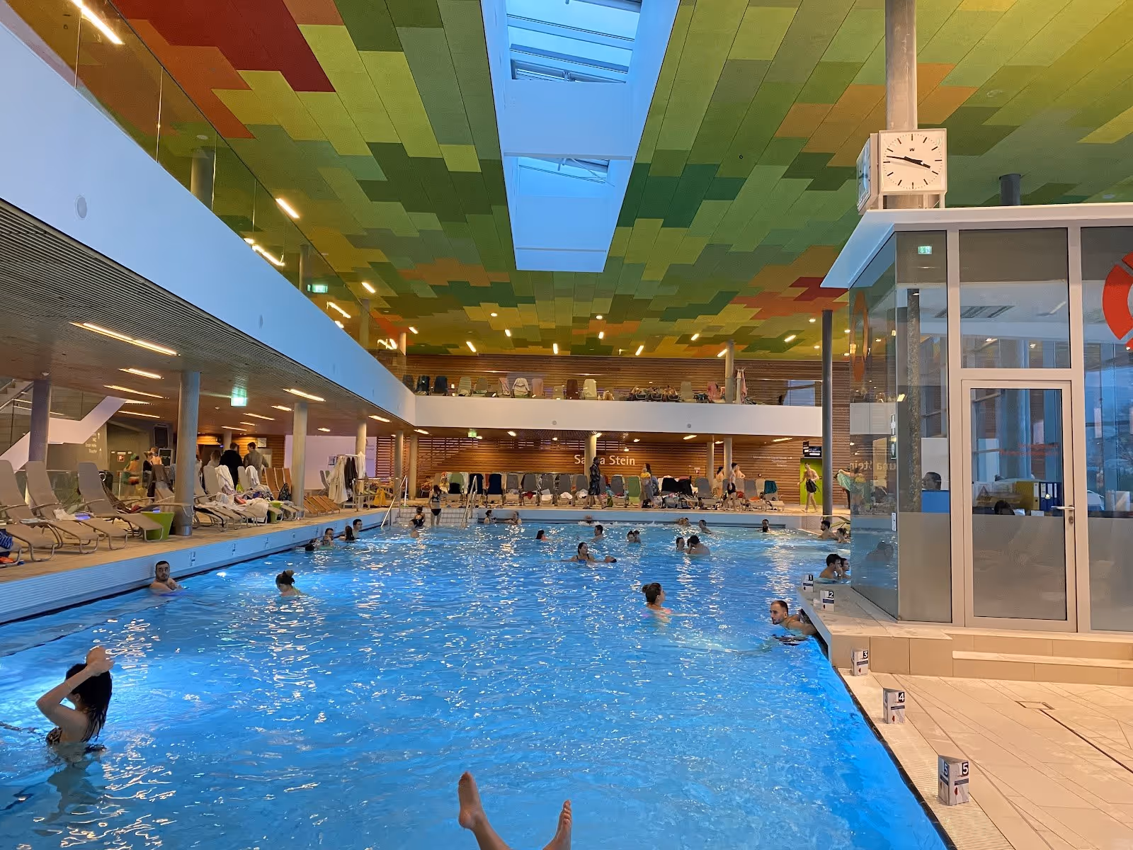 Indoor hot spring pool at Therme Wien in Vienna Austria with colorful ceiling panels and many people swimming and relaxing in the water.