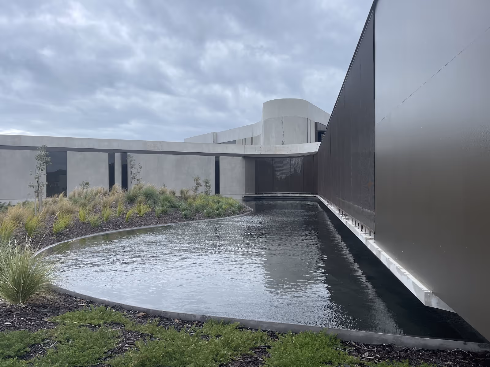 Long curved outdoor pool with dark water alongside modern gray building and landscaped garden at Alba Thermal Springs Australia.