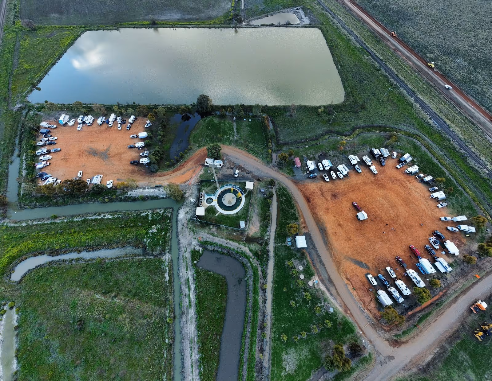 Aerial view showing the circular hot spring and surrounding parking area with many vehicles near green fields at Burren Junction Artesian B.