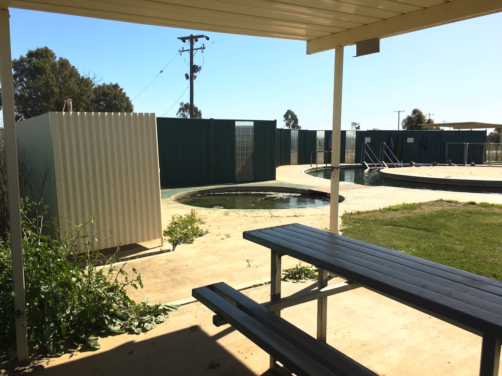 Shaded picnic table with outdoor seating next to multiple hot spring pools enclosed by green fencing at Burren Junction Artesian Bore Baths.