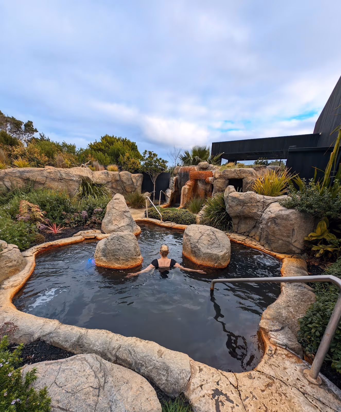 Woman soaking under a rocky cliff waterfall in an outdoor hot spring pool at Deep Blue Hot Springs near Warrnambool Australia.