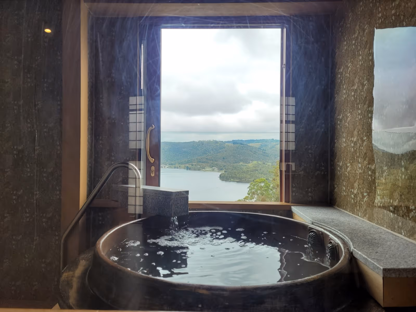 Indoor round wooden hot spring bath filled with clear water next to a large window showing a serene lake and green hills in South Bowenfels.