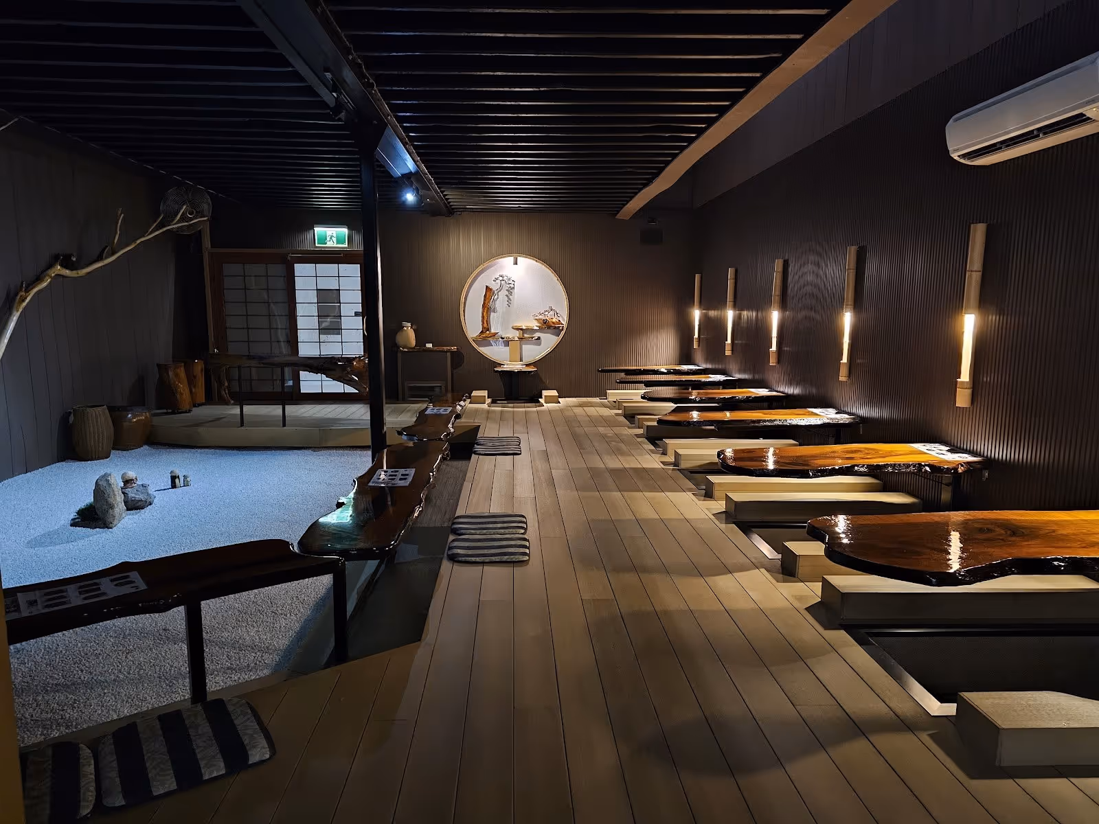 Interior of Japanese Bath House seating area with wooden tables over water foot baths, soft lighting, zen garden, and bamboo wall lamps in.