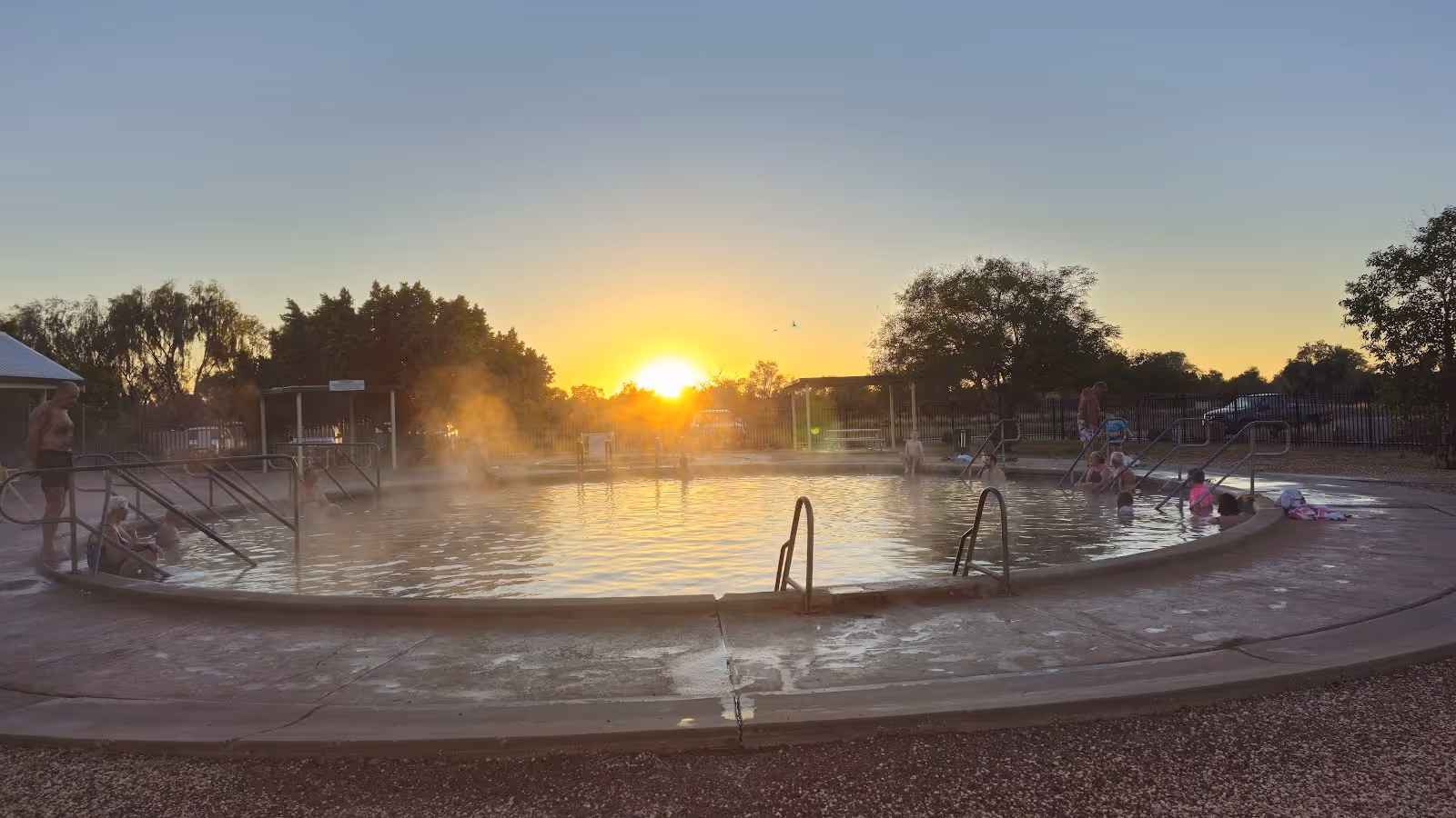 Steamy round hot spring pool with people at sunset under a clear sky at Lightning Ridge Artesian Bore Baths in New South Wales, Australia.