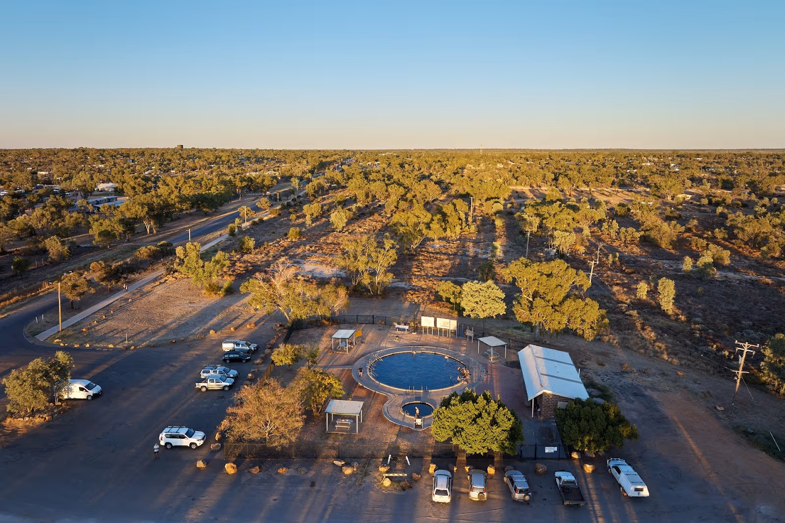 Aerial view of circular hot spring pool surrounded by trees and parked cars at Lightning Ridge Artesian Bore Baths in New South Wales, Aust.