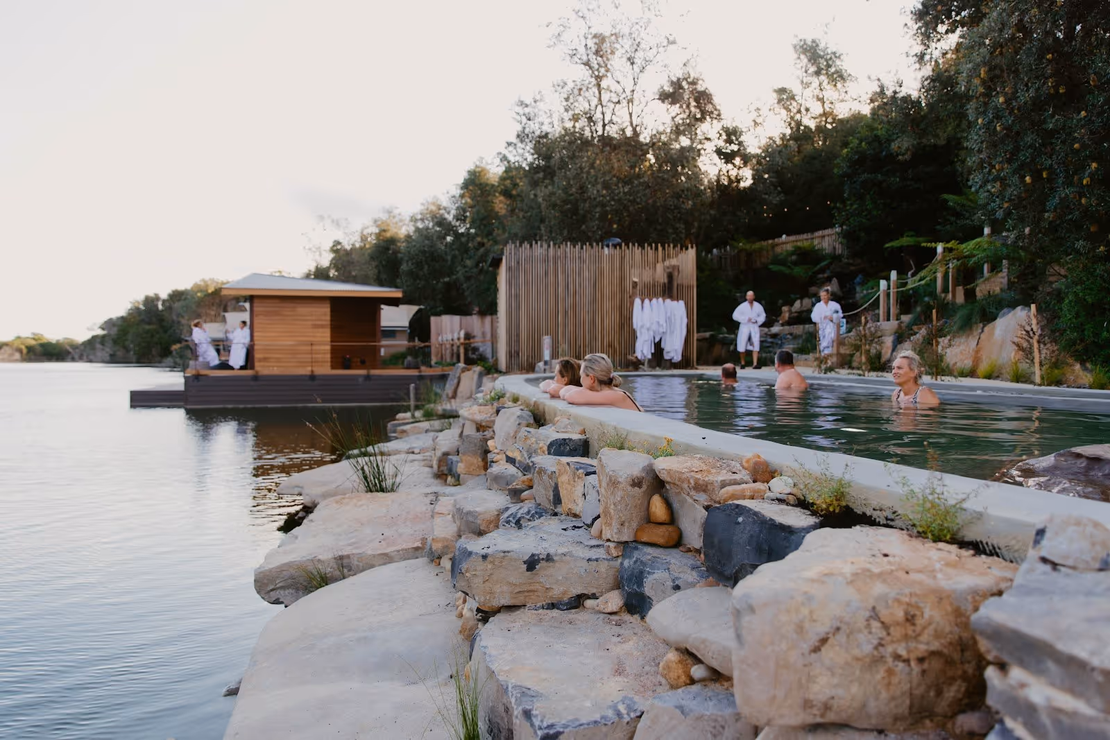 Several people enjoying a large outdoor mineral pool next to a rocky shoreline with robes hanging nearby at Metung Hot Springs, Australia.