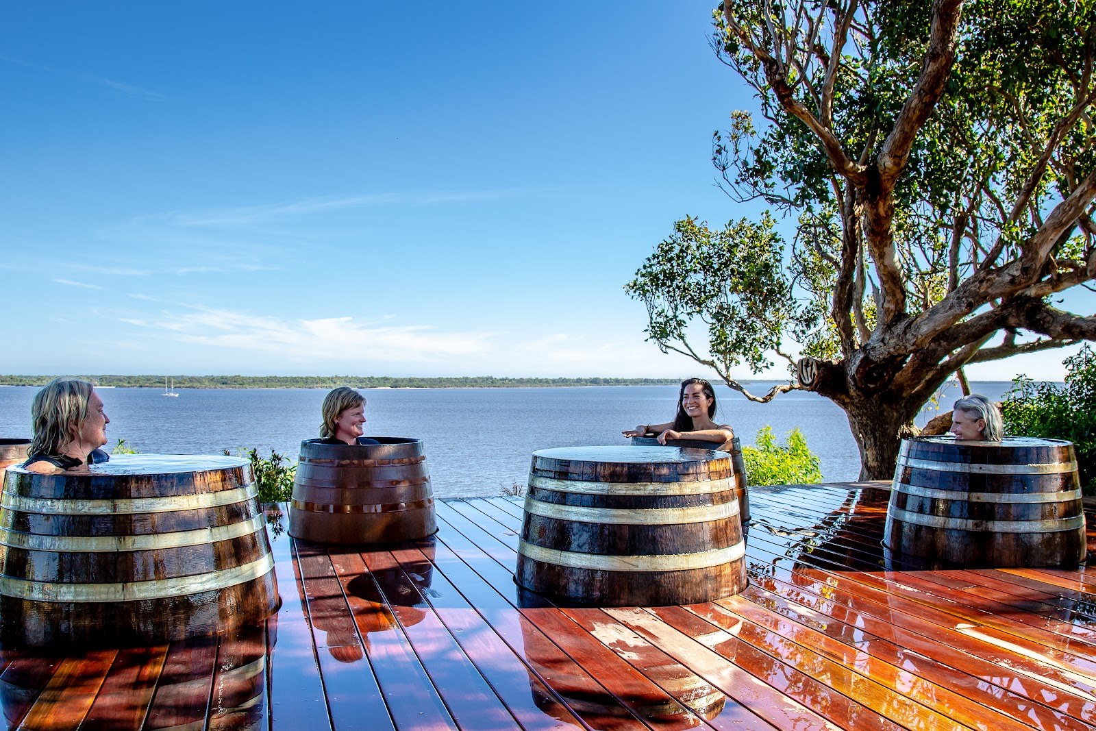 Four people relaxing in round wooden barrel spa pools on a polished deck with a tree and water view at Metung Hot Springs, Australia.