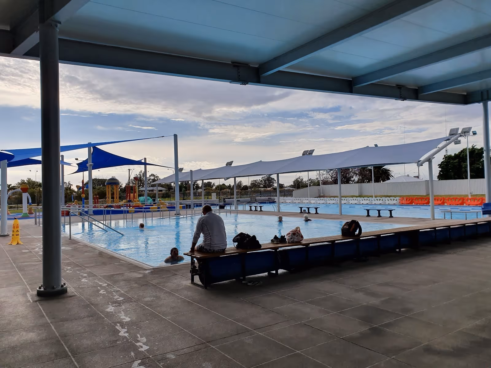 Covered outdoor pool area with people swimming and blue shade sails, benches along the pool edge near Moree in New South Wales.