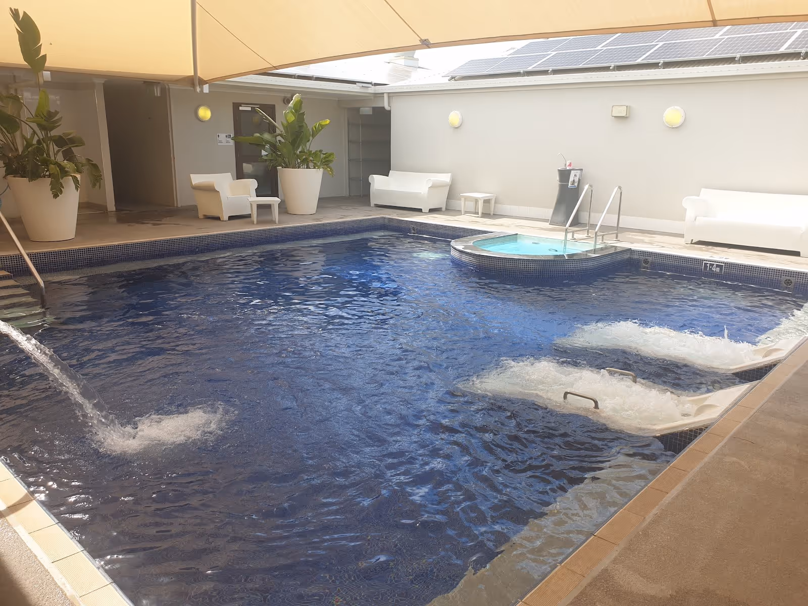 Indoor spa pool with dark blue tiles, clear water, white lounge chairs and large potted plants at Moree Artesian Aquatic Centre.
