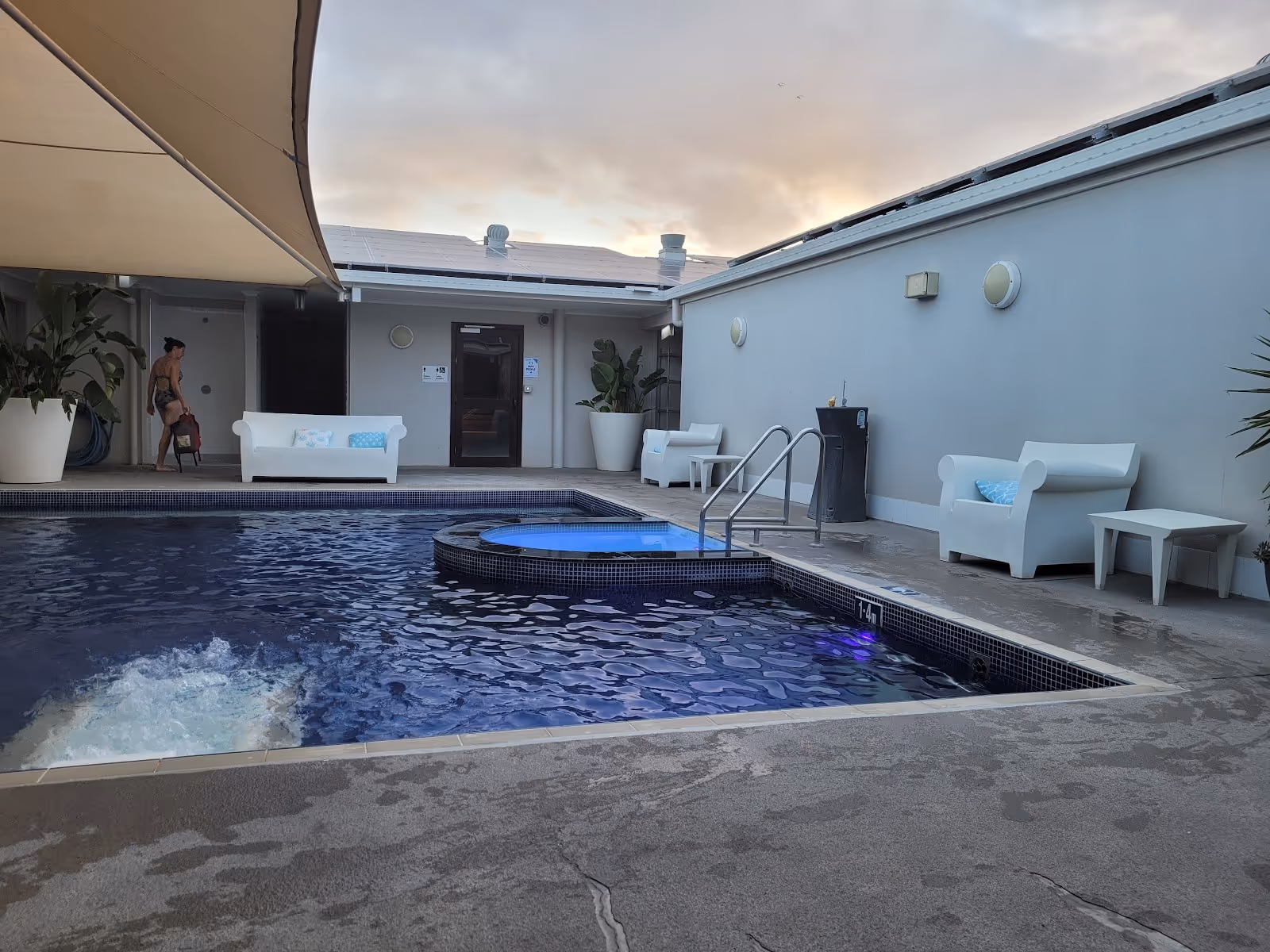 Evening light on indoor spa pool with dark blue mosaic tiles, bubbling water, white seating, and a person walking at Moree Artesian Aquatic.
