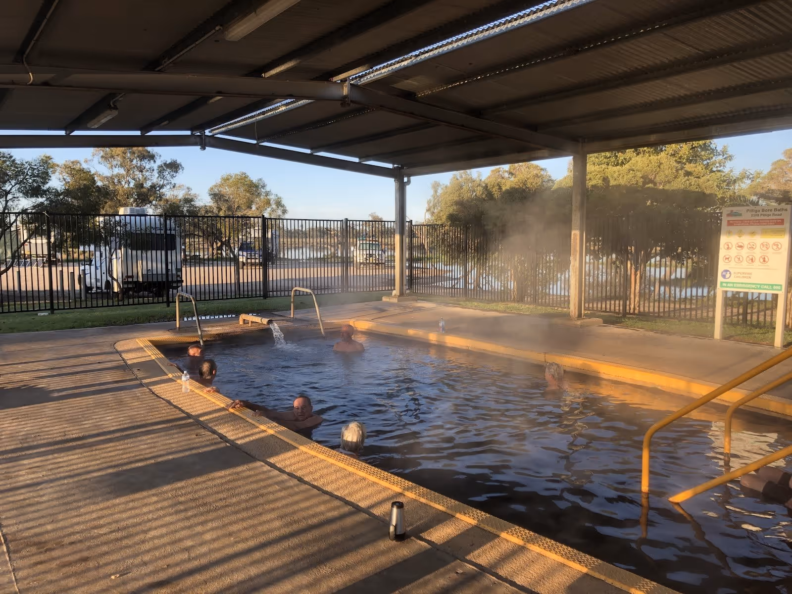 Covered outdoor pool with warm water and steam rising, several people relaxing inside at Pilliga Artesian Bore Baths near Pilliga NSW Austr.