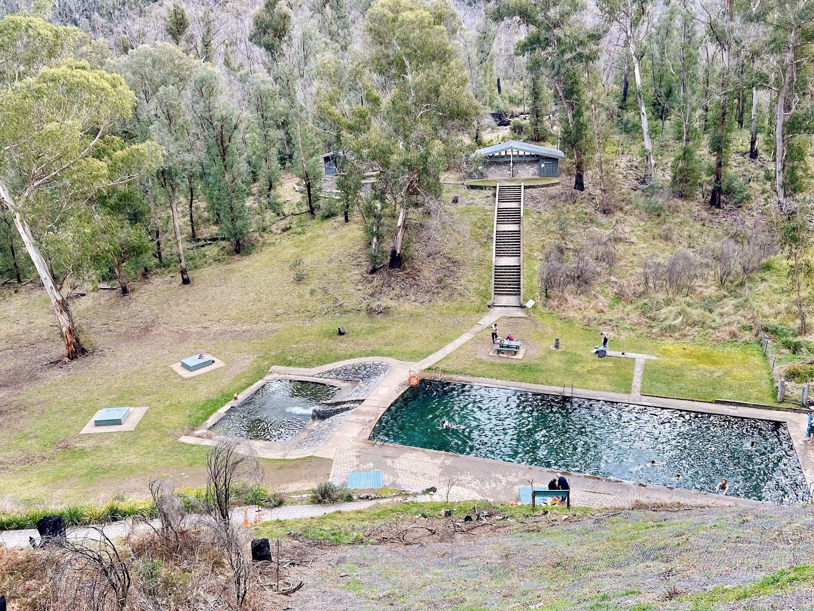 A rectangular outdoor thermal pool and a smaller pool sit in a grassy clearing with stairs leading up to a shelter at Yarrangobilly Caves t.
