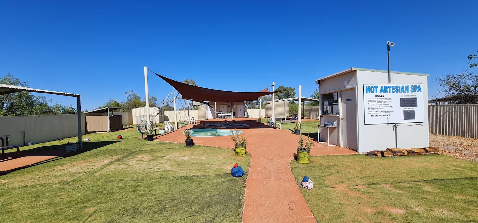 Wide view of Yowah Artesian Pools area showing multiple soaking pools surrounded by lawn and a shaded canopy on a sunny day.