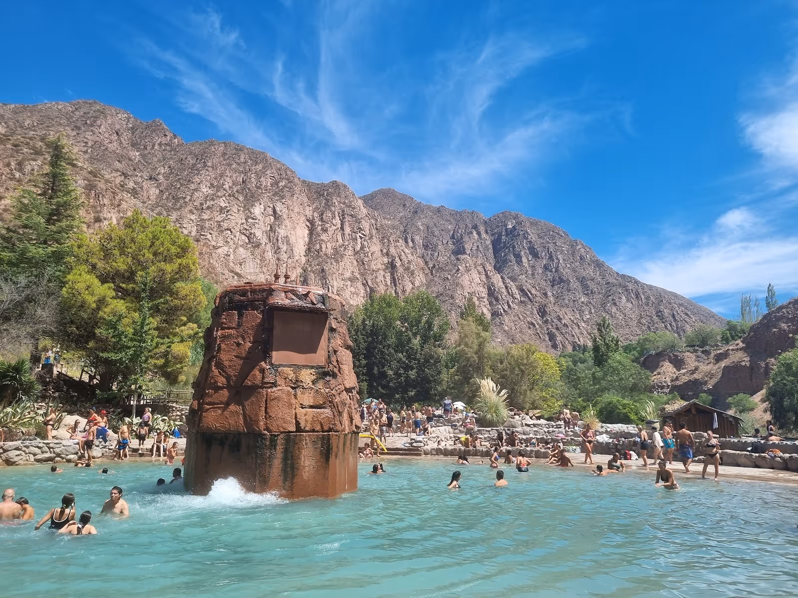 Large crowded hot spring pool at Termas de Cacheuta with a central stone water feature surrounded by rocky mountain landscape in Mendoza.