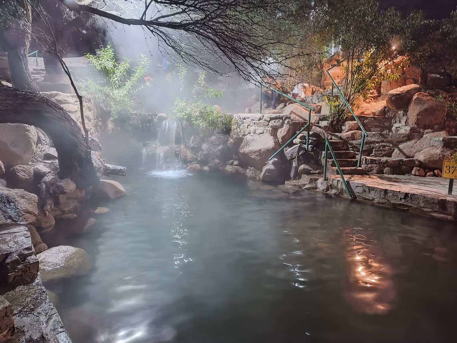 Steaming hot spring pool with natural rock edges and a stone staircase surrounded by trees at Termas de Fiambalá near Fiambalá, Argentina.