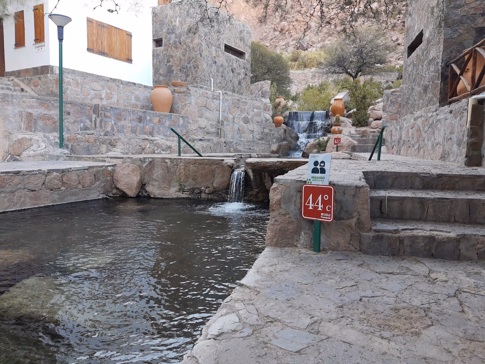 Stone pathways and multiple small hot spring pools with temperature signs and mountain cliffs visible at Termas de Fiambalá, Argentina.