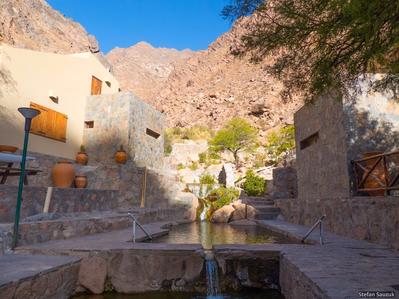 Hot spring pool bordered by stone walls and large clay pots under bright sunlight with mountains in the background in Fiambalá, Argentina.