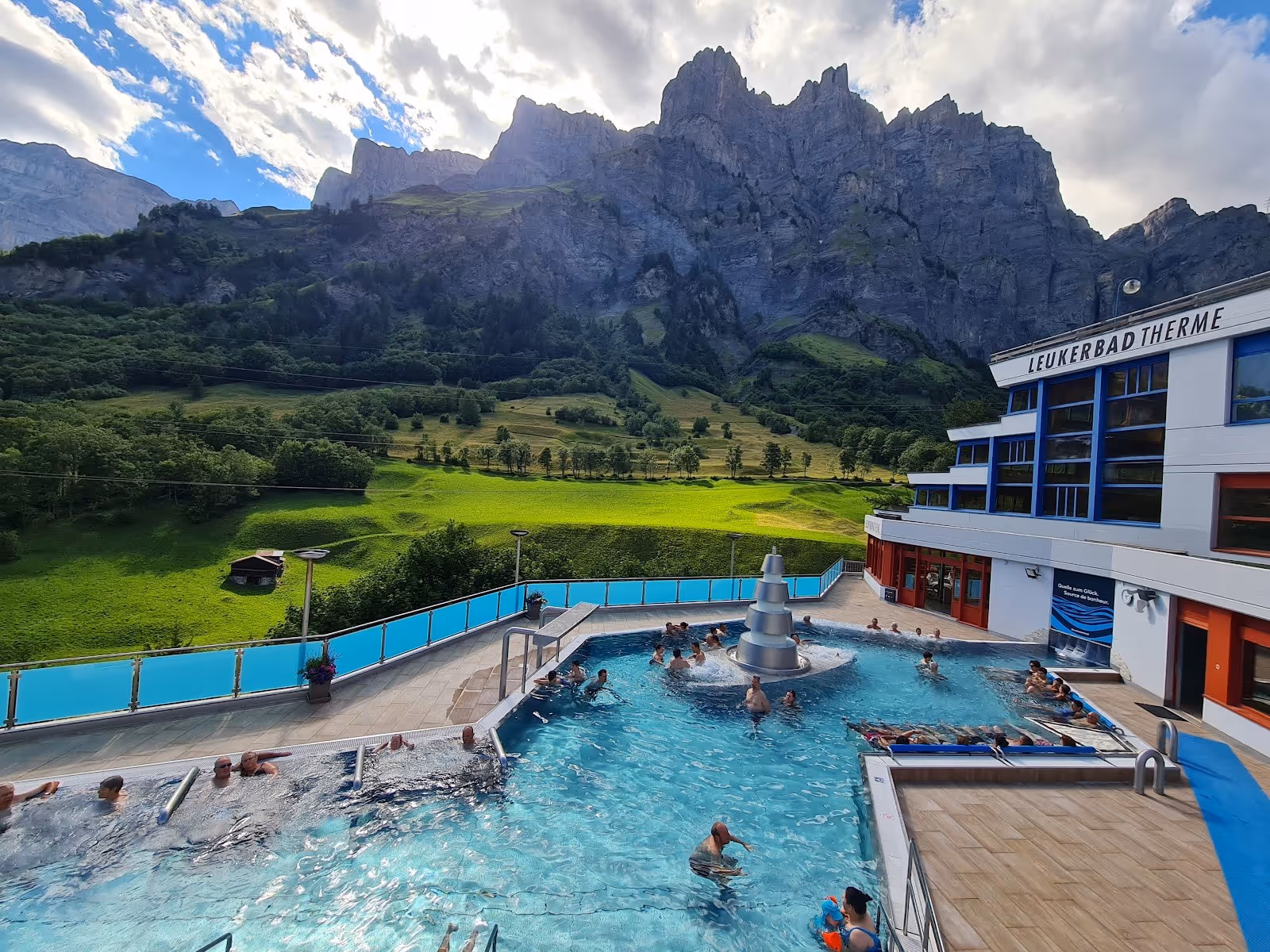 Crowded outdoor spa pool at Leukerbad Therme with people relaxing near a modern fountain under a partly cloudy sky and steep green mountain.