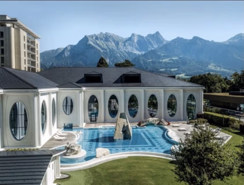 Daytime view of Tamina Therme outdoor mineral pool with blue tiles and triangular rock sculpture, hills in background at Bad Ragaz.