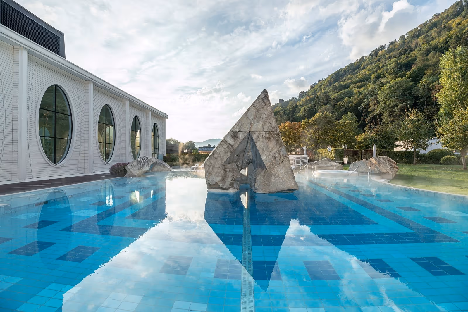 Steaming outdoor pool at Tamina Therme surrounded by green lawn and trees under cloudy sky near Bad Ragaz Switzerland.