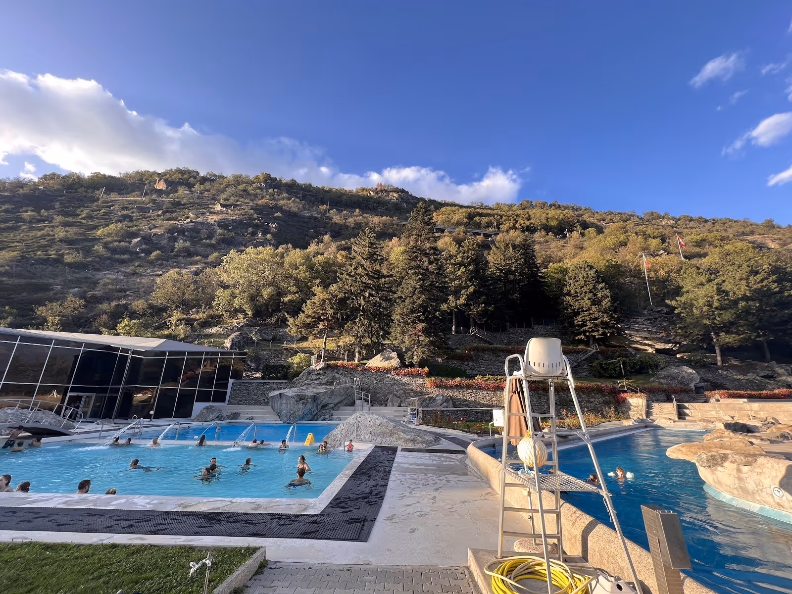 Outdoor soaking pools with bathers at Thermalbad Brigerbad with mountain hillside and scattered trees near Brig Switzerland.