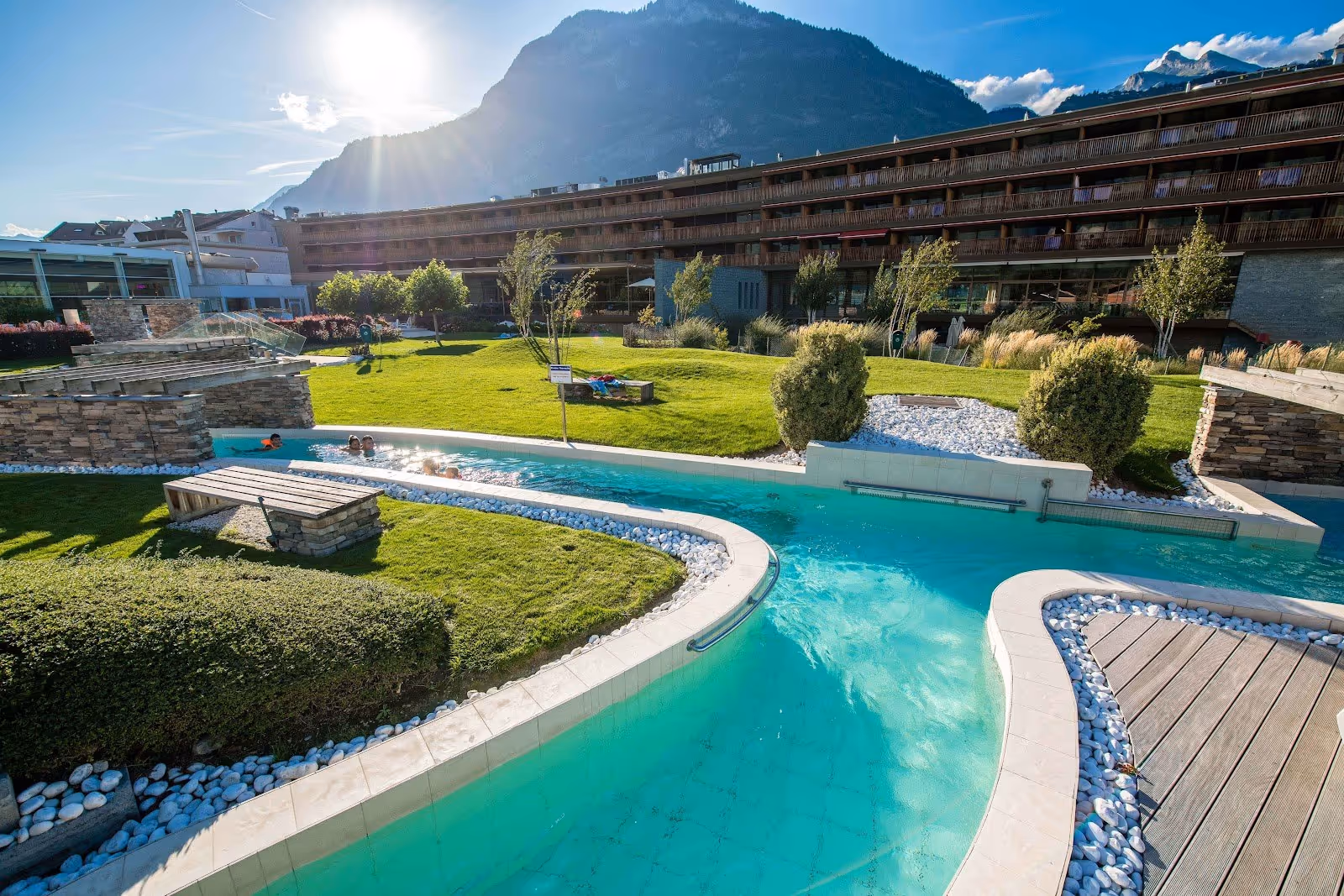 Sunlit mineral water pool with wooden benches and stone walls at Bains de Saillon near Martigny surrounded by grass and alpine peaks.
