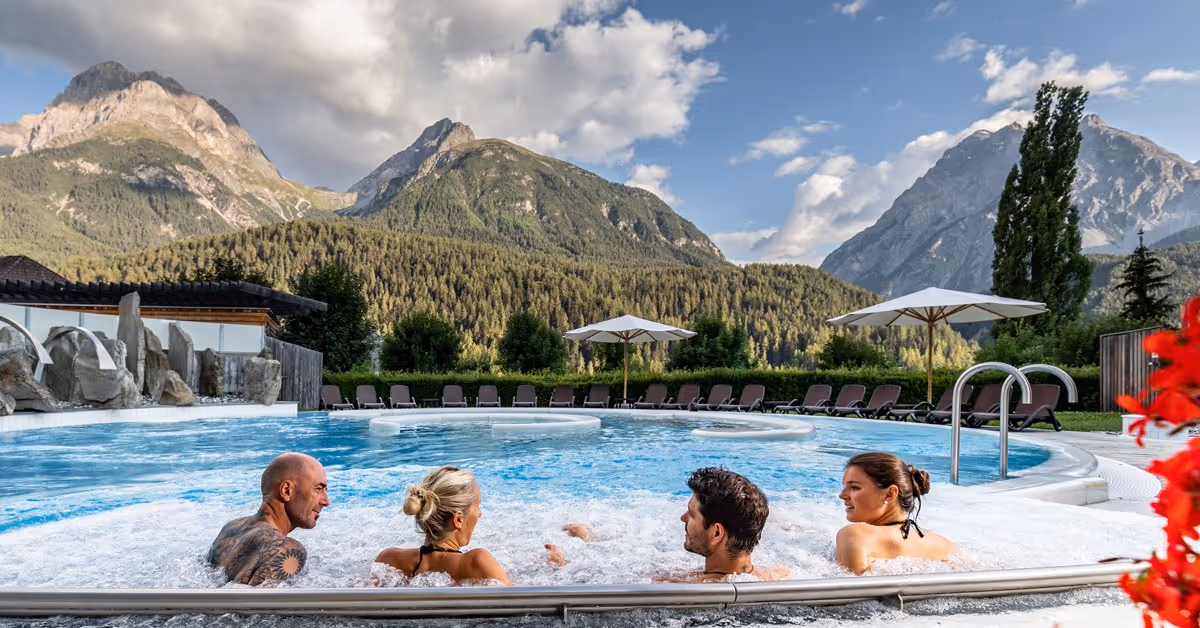 Four people relax in the outdoor bubbling hot spring pool at Bogn Engiadina Scuol with mountains and clouds in the background.
