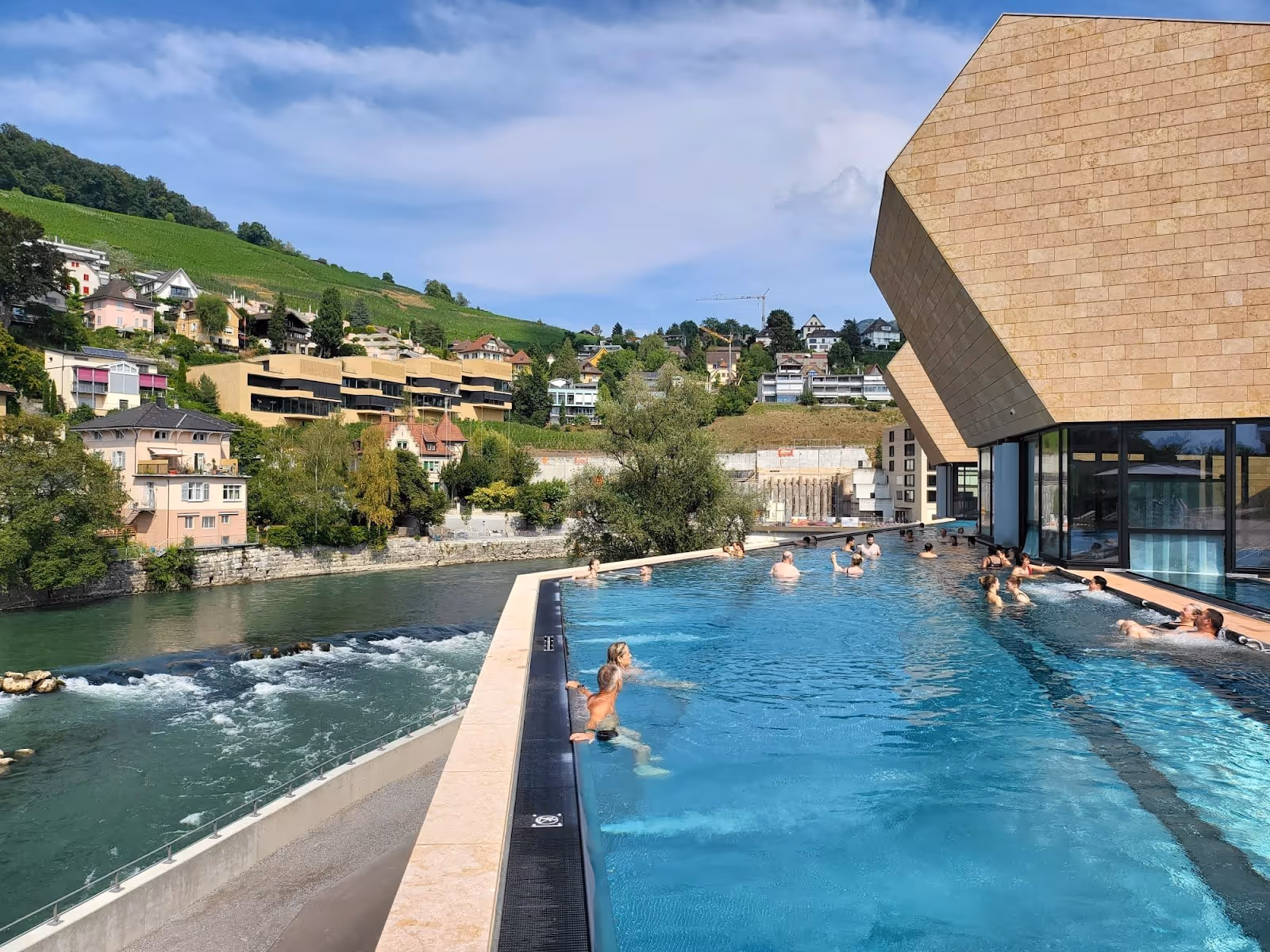 Outdoor pool at FORTYSEVEN Wellness-Therme with clear water and people relaxing near the building beside a river in Baden, Switzerland.