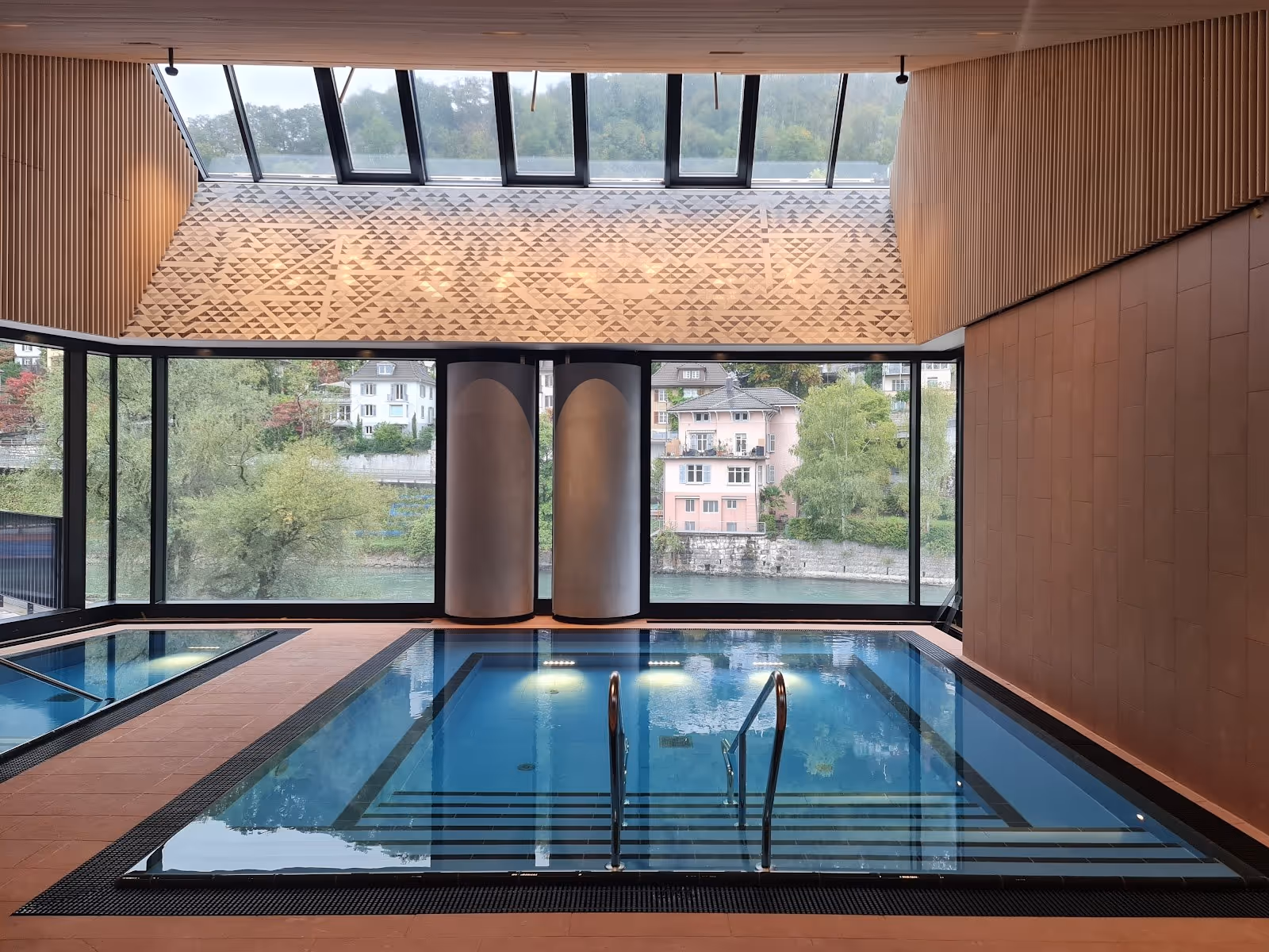 Indoor soaking pool with large windows overlooking a riverside view and modern wooden ceiling design at FORTYSEVEN Wellness-Therme in Baden.