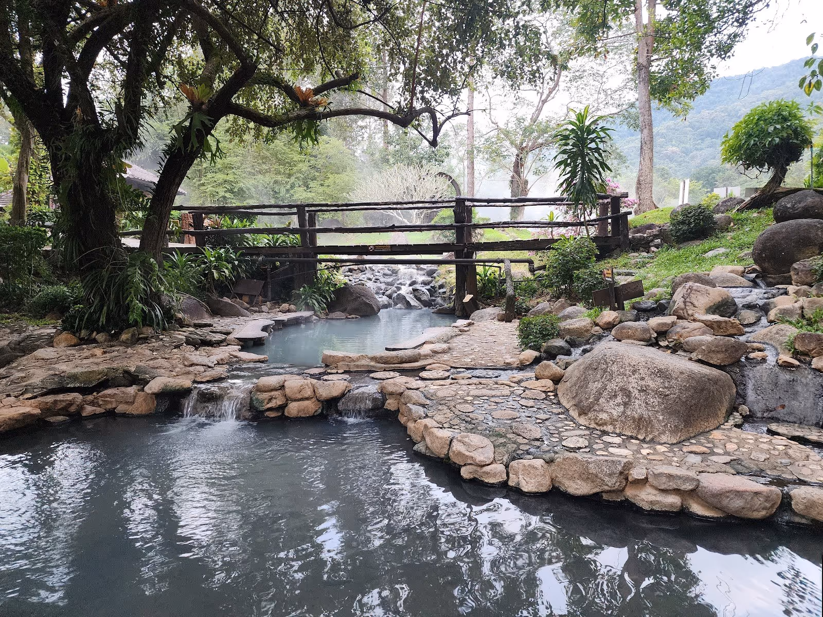 Outdoor hot spring pools at Chae Son Hot Springs in Nan, Thailand featuring stone walkways, a wooden bridge, and steaming mineral water.