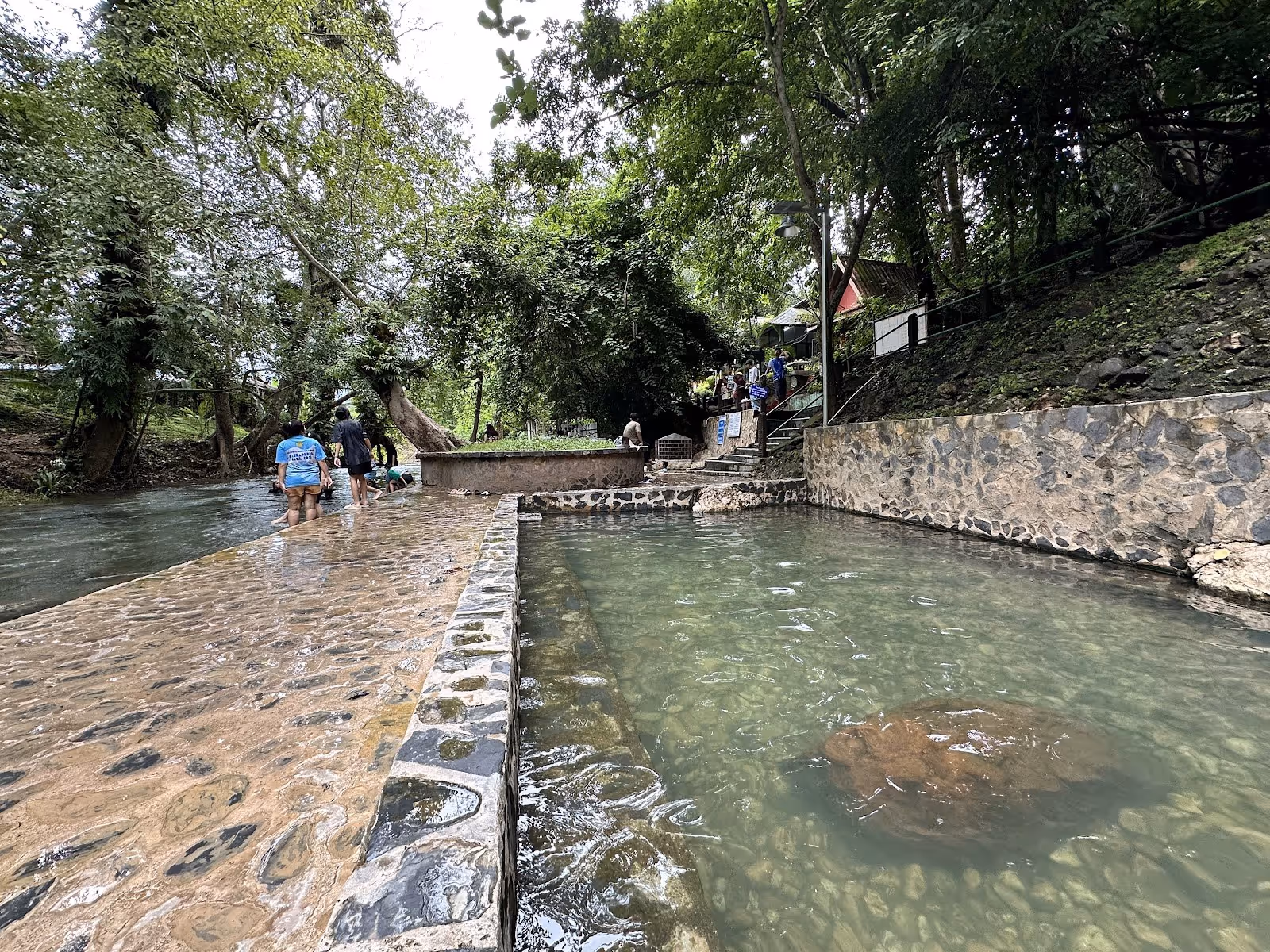 People walking beside clear water pools at Hin Dad Hot Spring with lush green trees in Thong Pha Phum, Nan, Thailand.